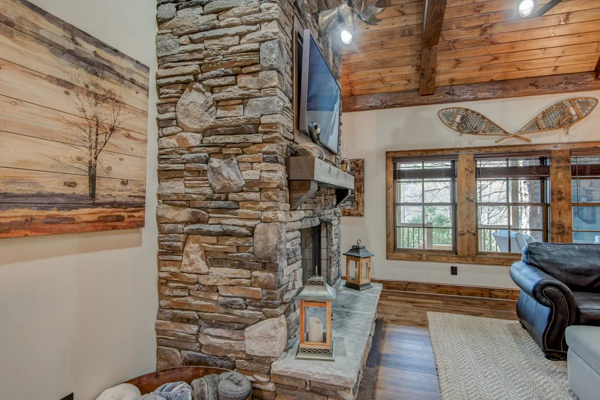 This rustic living area features a stone fireplace, wooden beams, large windows, and decorative lanterns. The comfortable seating and generous natural light provide a welcoming space for socializing and connecting with loved ones against a backdrop of scenic views.