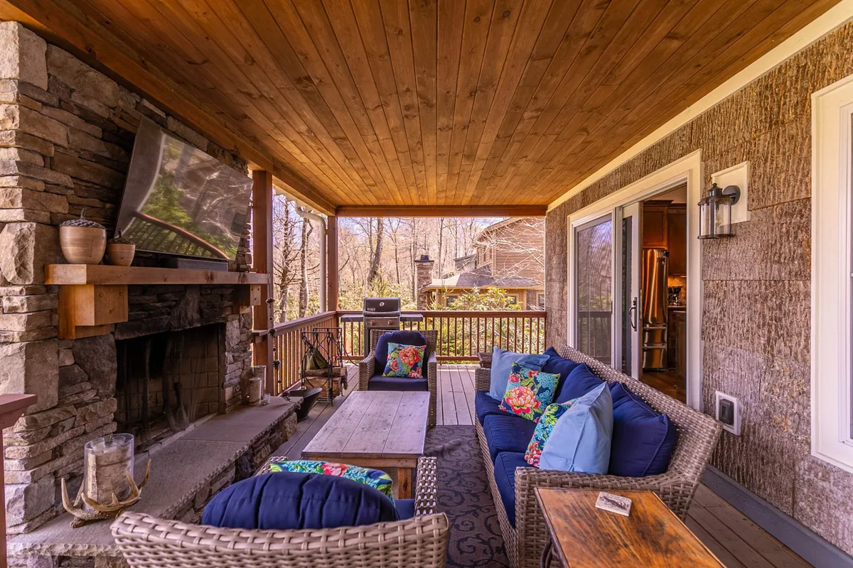This image highlights an elegant outdoor living area with a stone fireplace, comfortable seating, decorative pillows, and an outdoor grill. The cozy arrangement suggests a space for intimate gatherings, fireside conversations, and outdoor meals, all under the protection of a covered porch.