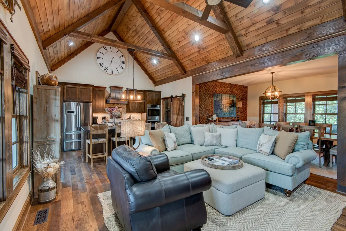 This image captures an inviting open-concept living and dining space, anchored by a large sectional sofa beneath wooden ceiling beams. The room connects seamlessly to a visible dining area, exuding warmth and togetherness, while a large window hints at views beyond, bringing in natural light.