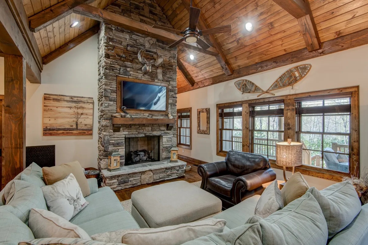 This image showcases the great room with a prominent floor-to-ceiling stone fireplace, a large plush sectional sofa, wooden beams, and a TV mounted above the fireplace. The expansive windows bring in abundant natural light and highlight the connection with the outdoors, while the cozy arrangement encourages relaxation and social gatherings.