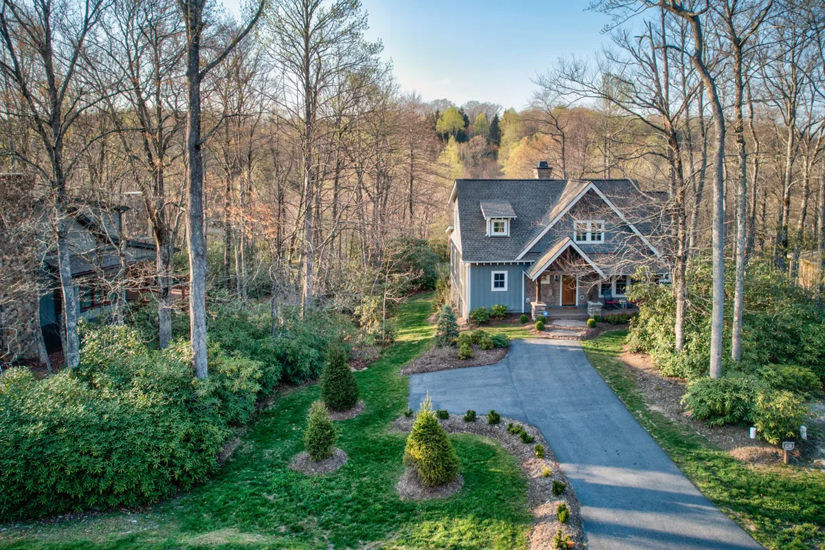 A wide exterior shot of the cottage, showing the home nestled among trees, with the driveway leading up and the porch visible. The tranquil surroundings evoke a sense of seclusion and immersion in nature, highlighting the property's connection to the outdoors.