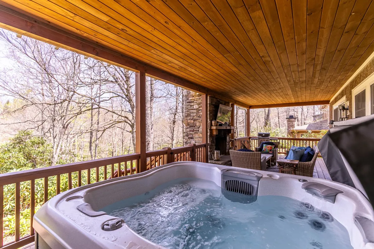 This image captures a tranquil scene of a hot tub outdoors, paired with a cozy seating area, natural forest surroundings, and an inviting fireplace. The setup is ideal for both solo relaxation and gatherings, highlighting the versatility and appeal of the porch area. Twilight lighting suggests it’s perfect for sunset or evening enjoyment.