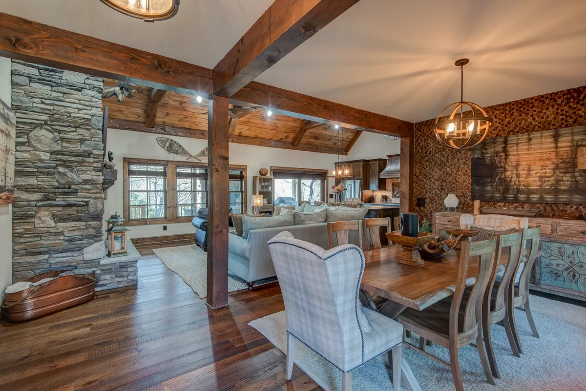 This interior shot blends the dining area and the great room, highlighting a stone fireplace, dining table, wooden beams, large windows with woodland views, and textured wall art. The communal layout encourages gatherings, whether for meals or relaxing by the fire.
