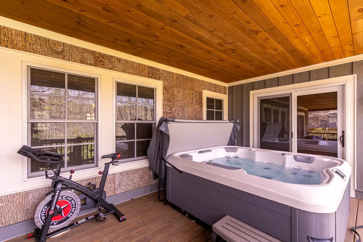A welcoming exterior scene featuring a hot tub nestled on a deck beside large windows, with an exercise bike for those who want to stay active. The natural outdoor setting offers a sense of tranquility, suggesting relaxation and wellness after a day of adventure.