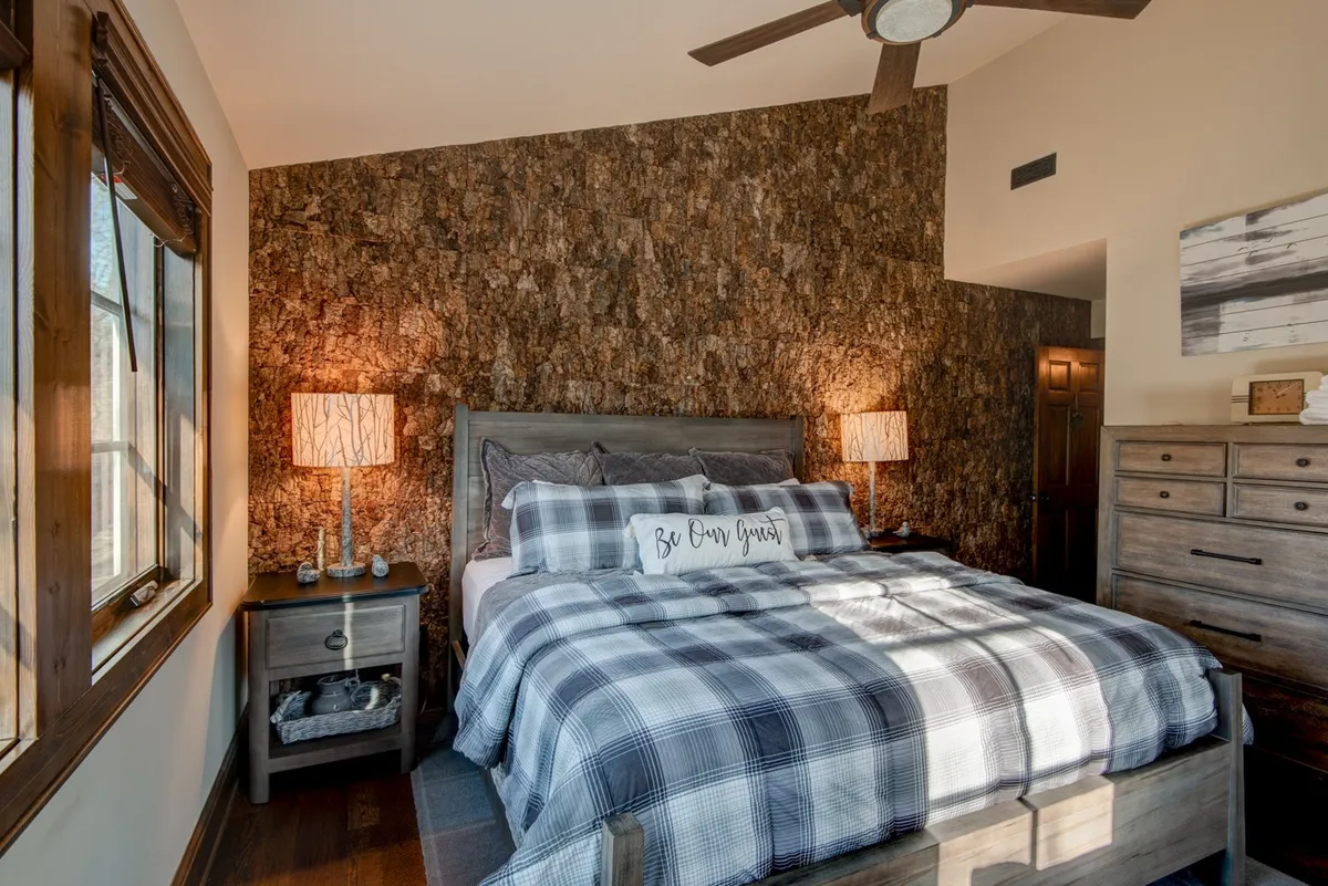 A spacious King bedroom suite featuring plaid bedding, a striking wood accent wall, and warm lighting from bedside lamps. Natural light fills the room, creating an inviting and cozy atmosphere ideal for relaxation. The rustic accent wall echoes mountain-inspired design, while the comfort-focused furnishings offer a sanctuary feel.