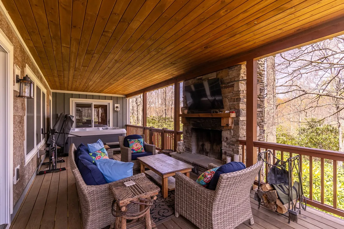 This image shows a covered back porch featuring comfortable seating, a bubbling hot tub, a cozy fireplace, decorative pillows, and a wooden ceiling. The warm ambiance invites relaxation and socializing, with views toward the surrounding trees enhancing the serene atmosphere. The space is obviously designed for year-round enjoyment, regardless of weather.