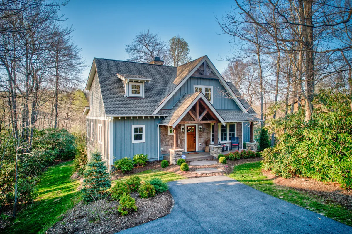 The image features the front porch of the cottage, complete with stone steps, a classic gable roof, and vibrant landscaping. The porch seating invites guests to relax and take in the natural beauty, offering an ideal spot for enjoying morning coffee or evening gatherings.