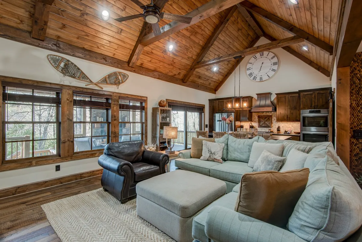 A spacious seating area with a large sectional sofa, wooden beams, and a prominent clock creates a cozy atmosphere designed for group relaxation. Large windows offer glimpses of the outdoors, suggesting easy access to nature and the wider community.